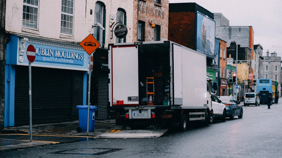 A white moving truck with its rear doors open is parked outside a commercial building on a city street during daytime, part of a home relocation process. The truck is positioned adjacent to the pavement, with the loading area visible, and a trolley loaded with boxes and packing materials inside. Nearby, a blue recycling bin is placed on the sidewalk. The building next to the truck has signage for 'Dublin Mouldings' with a partially visible window and a closed roller shutter door. Further along the street, other vehicles, including cars and a bus, are seen moving or parked, with additional storefronts and signs visible in the background. The scene depicts the logistics of furniture transport and packing, relevant to house removals and moving services, with natural daylight illuminating the urban environment.