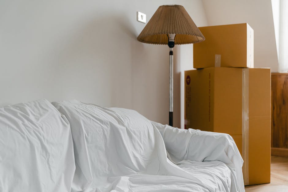 Inside a home's living room during a house removal, a white fabric-covered sofa is positioned in the foreground. Behind it, a tall, freestanding floor lamp with a beige pleated lampshade is located near the wall. To the right of the lamp, three stacked cardboard moving boxes are visible, with the largest at the bottom and the smallest at the top, arranged close to a wooden piece of furniture. The scene is illuminated with natural light, highlighting the room's neutral-toned walls and wooden flooring. The image captures part of the packing process with the boxes prepared for transport as part of a furniture transport or home relocation effort, consistent with the services offered by Man with Van Lisson Grove, a specialist in removals and moving logistics.