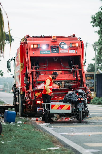 A worker wearing a red and yellow uniform is standing beside a large red commercial waste collection truck on a city street, with the truck's rear open and various debris on the ground nearby, including black plastic bags and loose rubbish. The worker appears to be handling or inspecting waste items during a bulky waste pickup process, with a municipal-style vehicle positioned on the roadside and the surrounding environment showing trees, street signage, and a building in the background. This scene illustrates the waste collection and logistics involved in home or business rubbish clearance, often part of house removals or relocation services provided by Man with Van Lisson Grove.