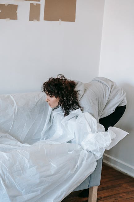 A woman with curly dark hair and light skin, dressed in a light grey hoodie, is leaning over a white sofa covered with white packing paper, inside a room with white walls. The wall behind her has four square patches of tape and paper, suggesting ongoing decoration or preparation for a home relocation or packing process. The wooden floor is visible beneath her, and the scene captures a moment during the packing or moving stage, reflecting the services offered by Man with Van Lisson Grove for residential removals and furniture transport.