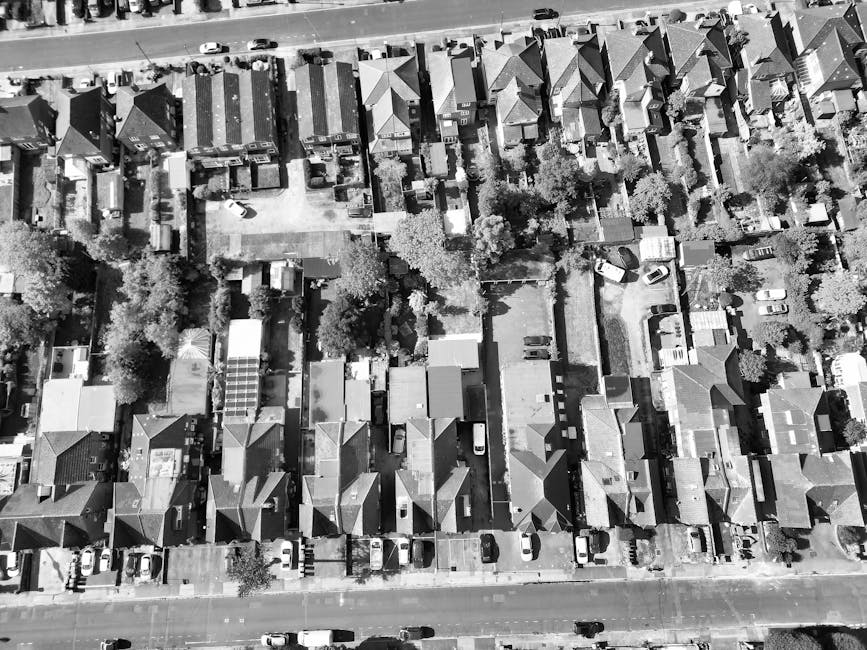 An aerial black-and-white photograph of a residential area showing rows of terraced houses with pitched roofs and small front gardens, trees lining the streets, and parked cars along the pavements. In the center, a moving van is parked on the street adjacent to a property, with open doors and a visible loading area on the pavement. Inside the van, several large cardboard boxes, wrapped furniture, and packing materials are arranged, ready for a house move. A man is seen carrying a large item wrapped in plastic and moving towards the van, while another person is near the doorway of a house, assisting with the placement of items. The scene captures a home relocation process involving furniture transport, packing, and loading, with the company Man with Van Lisson Grove providing professional moving services. The environment is well-lit by natural daylight, highlighting the detailed layout of the neighborhood and the logistics of moving household belongings.