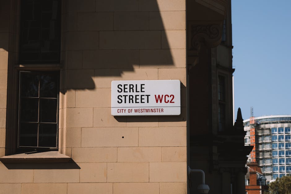 A street sign mounted on a beige stone building corner indicating 'Serle Street WC2, City of Westminster', with shadows cast on the wall above, and partially visible windows with black frames on the left. In the background, modern high-rise buildings and a clear blue sky are visible. The image captures the urban environment where home relocation or furniture transport services, such as those provided by Man with Van Lisson Grove, may operate during house removals or moving logistics within the Westminster area.