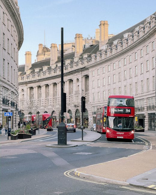 A busy city street corner with a large curved historic building featuring multiple chimneys and ornate architectural details in the background. On the roadway, there is a double-decker bus with a red exterior displaying route number 94 heading towards Edgware Green, partially visible as it moves past a pedestrian island. The street is lined with various vehicles, including a white van and parked cars, with road markings such as double yellow lines and pedestrian crossings clearly visible. Nearby, there are bus stops, street signs, and small planters with greenery, indicating an active urban environment typical of central London. In the foreground, part of a wide pavement and a lamppost are visible, with people walking along the sidewalk. The scene captures a moment of everyday city life, relevant to local moving and transportation logistics facilitated by companies like Man with Van Lisson Grove.
