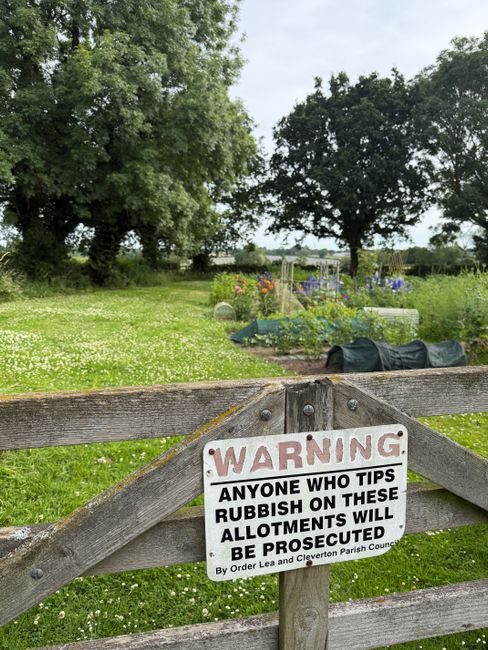 A weathered wooden gate with a warning sign attached, warning that tipping rubbish on these allotments will lead to prosecution, as issued by the Cleverton Parish Council. In the background, there is a lush, green outdoor area with large trees and open grassy space, some garden beds with colorful flowers, and gardening tools and equipment such as black covered containers, a wheelbarrow, and a small greenhouse or plant support structure. The sky is overcast, providing soft natural lighting that highlights the rural setting, which appears to be part of a community allotment or garden area. This outdoor scene is unrelated to house removals but focuses on local community regulations and outdoor environment, with the natural landscape and gardening materials in view.