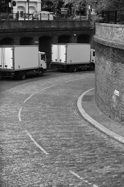 A black and white photograph of a cobbled street at a loading bay area next to a brick wall with an arched bridge above. Two white box trucks are parked side by side, parallel to the brick wall, engaged in the loading process. The trucks are positioned on the street, with one facing towards the bridge and the other facing away. The street features marked dashed white lines indicating parking or loading zones. Behind the trucks, the arched bridge structure is visible, supporting a balcony with fencing and potted plants above. The scene appears to be part of a residential or commercial back area used for home relocation or furniture transport, with the trucks likely used by a removals service such as Man with Van Lisson Grove to perform a house move or delivery. The environment is lit naturally, with clear visibility of all objects involved in the loading or unloading activity.