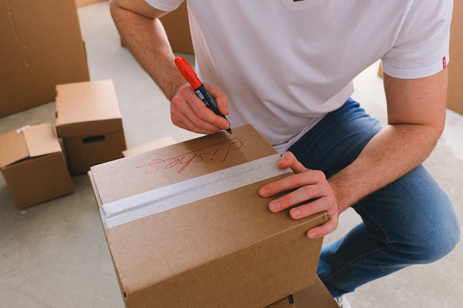 A person wearing a white t-shirt and blue jeans is kneeling on the floor inside a property, preparing to label a cardboard box with a red marker. The box is sealed with two strips of white packing tape across the top, and features the handwritten label 'SILAS' in red ink. Several other cardboard boxes of various sizes are stacked nearby on the floor, some with cut-out handles. The room appears to be part of a home or office, with plain walls and a light-colored floor, possibly concrete. Visible in the background are additional boxes and packing supplies, indicating an ongoing packing or home relocation process. The image captures the loading and packing stages of furniture transport or house removals, with the focus on careful labelling for organized moving logistics, as used by companies like Man with Van Lisson Grove.