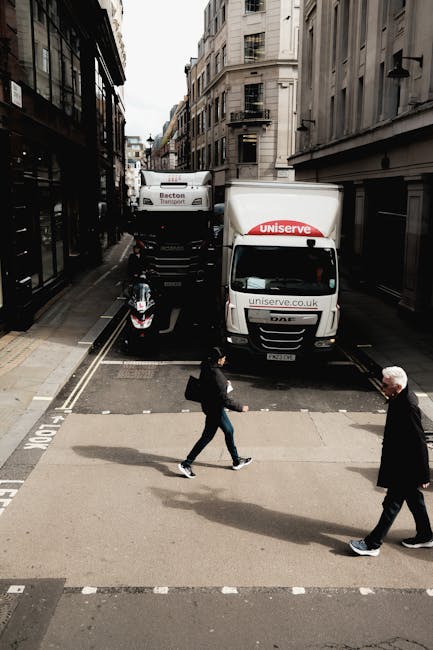 A city street scene showing a narrow road with tall, multi-story buildings on either side, featuring residential and commercial architecture. A white MAN delivery van with 'uniserve' branding on the front and side is parked partially in the middle of the street, behind which a larger truck with 'Becton Transport' branding is positioned, indicating loading or unloading activity. A black scooter is parked beside the vehicles, close to the curb. In the foreground, a woman wearing a dark jacket and jeans is crossing the street while carrying a bag, and an older man with white hair dressed in dark clothing is walking in the opposite direction on the crosswalk. The scene is illuminated by natural daylight, capturing a typical urban movement situation related to house removals or furniture transport, with the vehicles and pedestrian activity suggesting ongoing logistic or home relocation processes. Details such as the vehicles, packaging materials, and street environment highlight aspects relevant to moving services by Man with Van Lisson Grove.