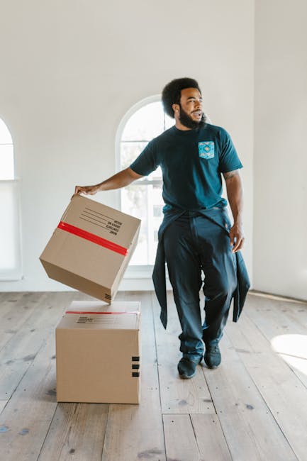 A man with dark curly hair and a beard, dressed in a navy blue t-shirt and dark trousers, is indoors in a well-lit room with white walls, wooden flooring, and large arched windows allowing natural light. He is holding a cardboard box wrapped with red packing tape in his right hand, preparing to move or load it. In front of him, there is another closed cardboard box on the floor, which is also likely part of a home relocation process. Behind him, the room appears empty, suggesting it may be a staging area for furniture transport or packing. This scene illustrates furniture transport and packing during house removals, with Man with Van Lisson Grove providing professional moving services, ensuring efficient and careful handling of items during a house move.
