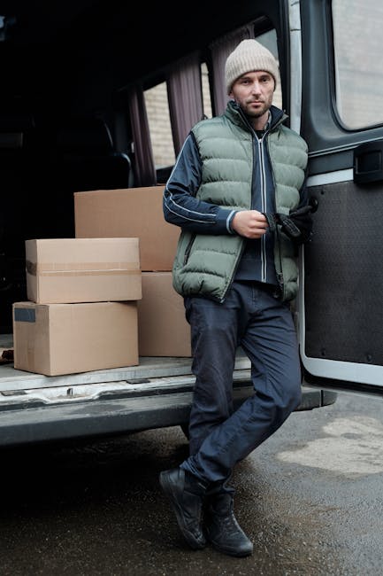 A man with a serious expression, wearing a light grey knit beanie and a padded green and navy jacket, stands inside a van with a dark interior. Behind him, there are cardboard boxes stacked on a troll